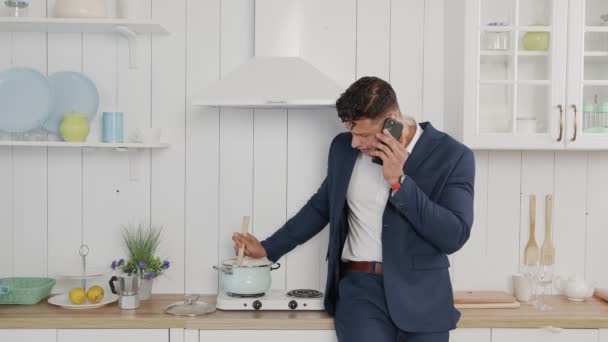 Чоловік у синьому діловому костюмі говорить по телефону і готує вечерю — стокове відео