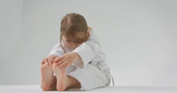 Het meisje zit op de vloer met haar benen recht en trekt haar vingers naar haar tenen. — Stockvideo