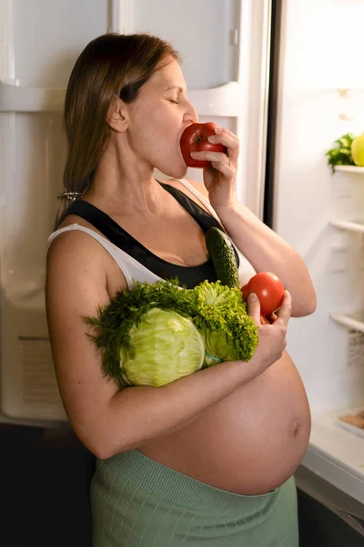 Těhotná žena drží čerstvé ovoce v blízkosti ledničky. Žena jí červené jablko. — Stock fotografie