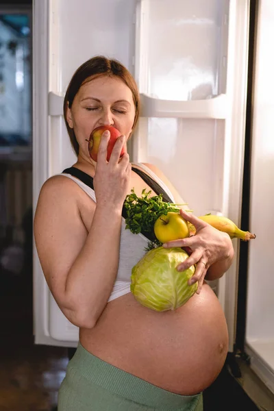 Mujer embarazada sosteniendo frutas frescas cerca del refrigerador. Mujer comiendo una manzana roja. —  Fotos de Stock