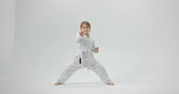 La chica practica karate en casa. El niño está vestido con un kimono sobre un fondo blanco.. — Vídeo de stock