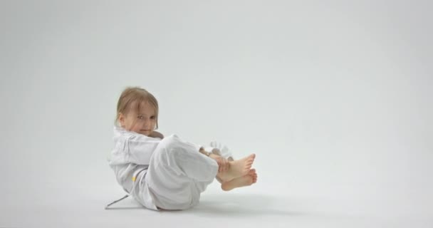 Het kind gaat op de grond zitten en probeert zijn benen op te tillen. Het meisje in een kimono doet sportoefeningen. — Stockvideo