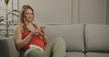 Hamile kadın hamile fotoğrafları için fikirlere bakıyor. Dikkatlice telefona bakıyor ve gülümsüyor..