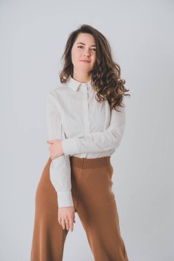 Beyaz arka planda genç bir kız kameraya bakar. Kız kahverengi pantolon ve beyaz gömlek giymiş..