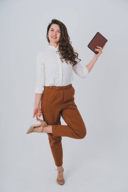 Kız bir bacağının üzerinde duruyor, diğerini bükülmüş bir kolla tutuyor. Elinde bir defterle, kameraya gülümsüyor..