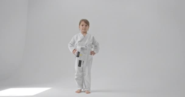 L'enfant concentré regarde la caméra, la frappe et la jette au sol. Bébé en kimono blanc sur fond blanc. — Video