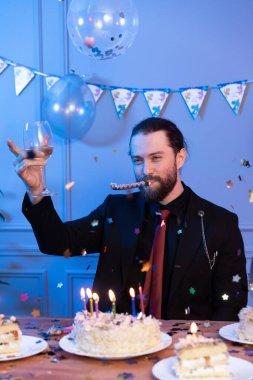 A man raises a glass of champagne for his birthday. He is sitting at the table without friends because he does not have them. In the air confetti and on the table a cake with candles.