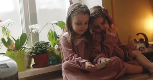 Aynı pembe elbiseli küçük kızlar annelerinin telefonunda eğitim oyunları oynuyorlar. Kızlar yan yana oturmuş telefona bakıyorlar.. — Stok video
