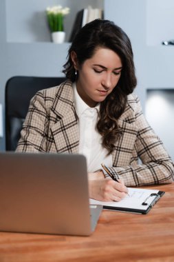 Bilgisayarda uzaktan çalışan ve kağıda not alan bir öğrenci. Odaklanmış bakış. İş kıyafetleri.