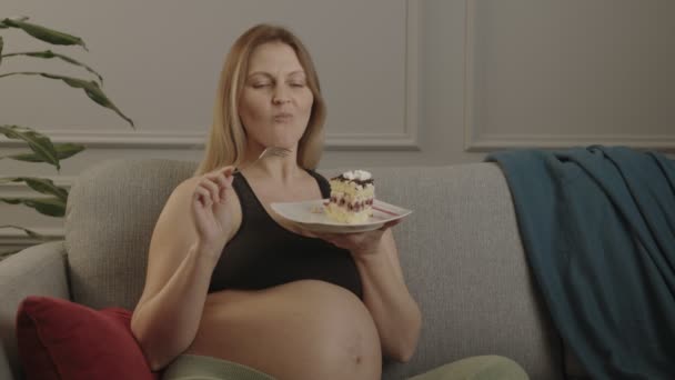 Mujer embarazada feliz comiendo un pedazo de pastel sentado en el sofá. — Vídeos de Stock