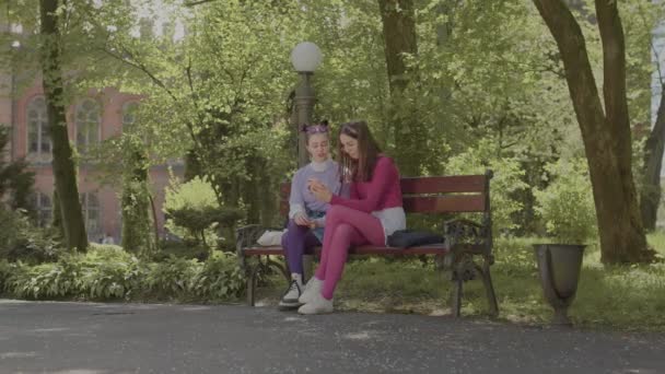 Les copines s'assoient sur un banc dans le jardin et regardent des vidéos au téléphone. — Video