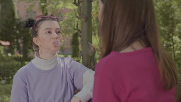 Meisjes spelen met kauwgom op een bankje in het park. De meisjes hebben plezier die een grotere zeepbel opblaast. — Stockvideo