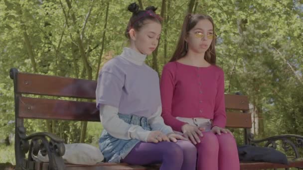 Két tinédzser lány ül egy padon komoly arccal. Szégyen megérinteni egymást.. — Stock videók