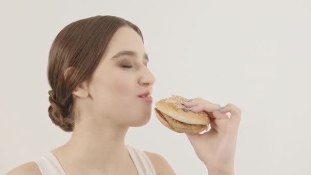 A menina deliciosamente come um hambúrguer em um fundo branco. A menina come junk food. — Vídeo de Stock