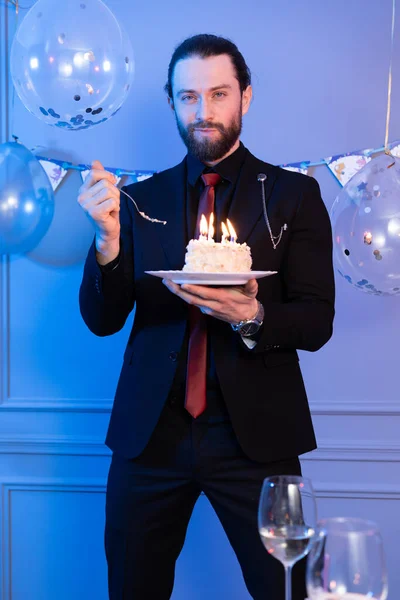 A man holds a cake with a fork and looks at the camera. Candles are burning on the cake.