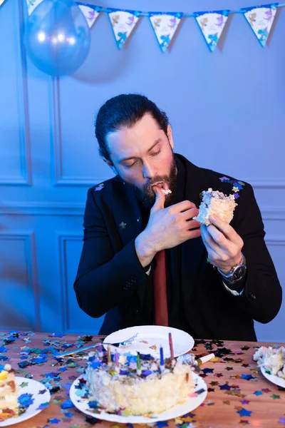 O homem está sentado à mesa segurando um pedaço de bolo e olhando para ele. Celebração de aniversário atípica. — Fotografia de Stock