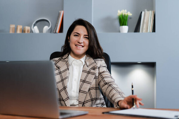 The girl is sitting at the table and smiling. Happy girl working as a freelancer on a laptop.