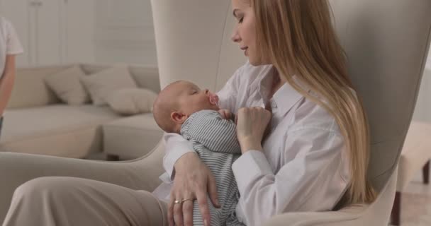 Mama sitzt auf einem Stuhl und hält ein Baby in der Hand. Im Hintergrund rennt das Kind durch den Raum. — Stockvideo