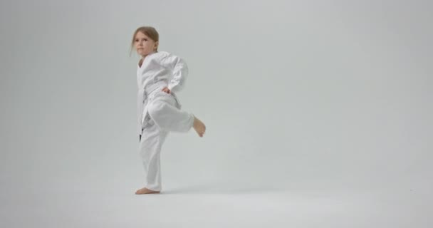 Het kind staat op zijn voeten met zijn handen op zijn riem en gooit een been schop. Het kind voert karate technieken uit. — Stockvideo