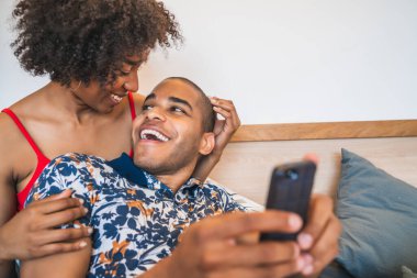 Evde cep telefonuyla selfie çeken mutlu genç Latin çiftin portresi. Yaşam tarzı ve ilişki kavramı.