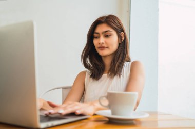 Bir kafede otururken dizüstü bilgisayarını kullanan genç Latin kadının portresi. Teknoloji ve yaşam tarzı kavramı.