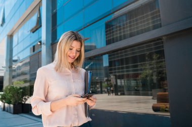 Ofis binalarının önünde beklerken cep telefonunu kullanan genç bir iş kadınının portresi. İş ve başarı kavramı.