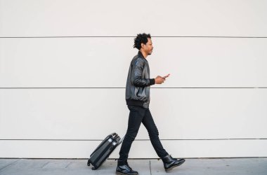 Turist adam cep telefonunu kullanıyor ve dışarıda bavulunu taşıyor..