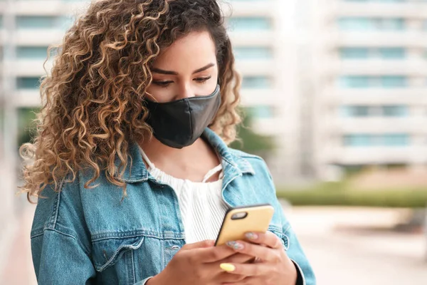 Latin woman wearing a face mask while using her mobile phone outdoors.