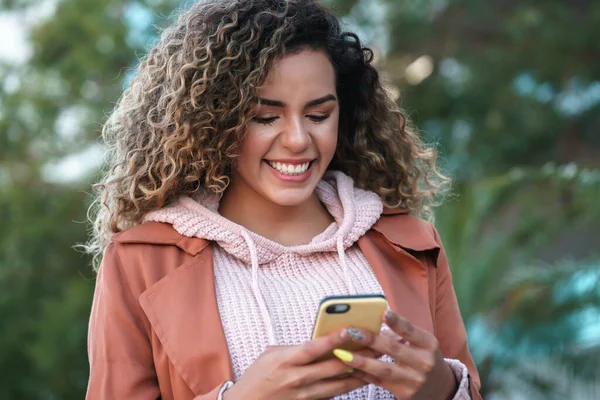 Mujer latina sonriendo mientras usa un teléfono móvil al aire libre en la calle. —  Fotos de Stock