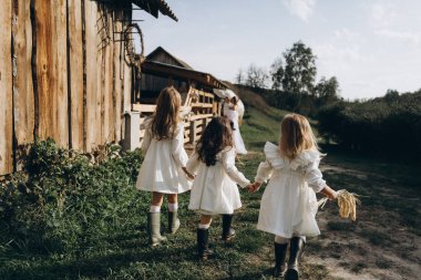 Beyaz bohem bir elbise giymiş güzel bir kadın çocuklarıyla ahırın yanında hayvanlarla dikiliyor. 