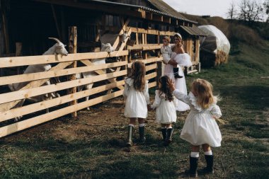 Beyaz bohem bir elbise giymiş güzel bir kadın çocuklarıyla ahırın yanında hayvanlarla dikiliyor. 