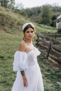 Beyaz bohem elbiseli güzel bir kadın çayırda yürüyor ve kameraya poz veriyor. 
