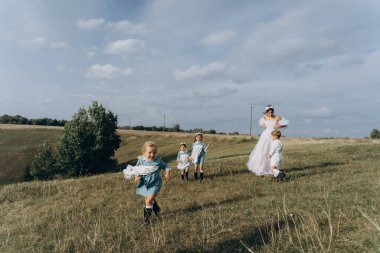Beyaz bohem elbiseli güzel bir kadın çocuklarıyla kırsalda yürüyor. 