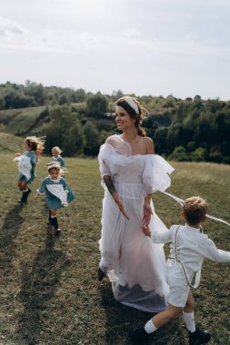 Beyaz bohem elbiseli güzel bir kadın çocuklarıyla kırsalda yürüyor. 