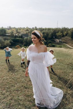 Beyaz bohem elbiseli güzel bir kadın çocuklarıyla kırsalda yürüyor. 