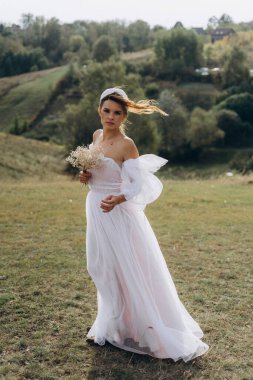 Beyaz bohem elbiseli güzel bir kadın çayırda yürüyor ve kameraya poz veriyor. 