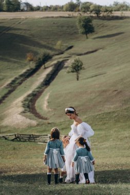 Beyaz bohem elbiseli güzel bir kadın çocuklarıyla kırsalda yürüyor. 