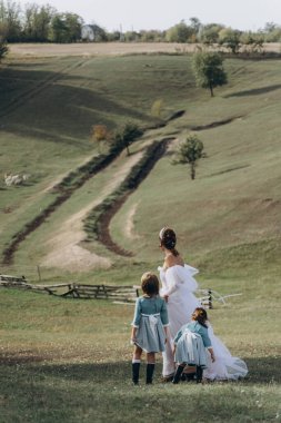 Beyaz bohem elbiseli güzel bir kadın çocuklarıyla kırsalda yürüyor. 