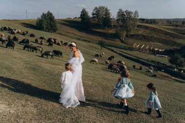 Beyaz bohem elbiseli güzel bir kadın çocuklarıyla kırsalda yürüyor. 