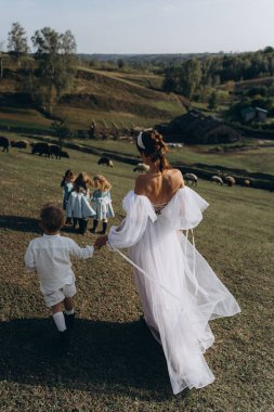 Beyaz bohem elbiseli güzel bir kadın çocuklarıyla kırsalda yürüyor. 