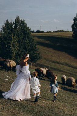 Beyaz bohem elbiseli güzel bir kadın çocuklarıyla kırsalda yürüyor. 