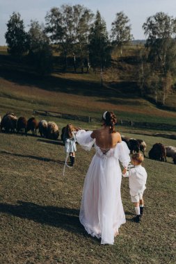 Beyaz bohem elbiseli güzel bir kadın çocuklarıyla kırsalda yürüyor. 