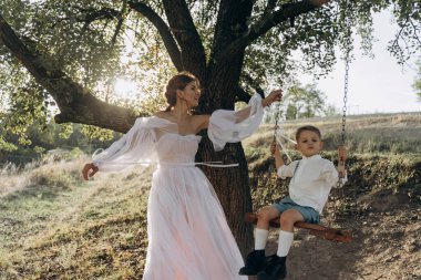 Beyaz bohem elbiseli güzel anne oğlunu sallıyor kırsal bir ağaçta salıncakta 