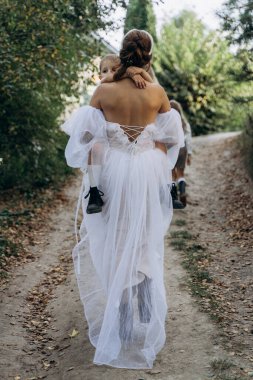 Beyaz bohem bir elbise giymiş güzel bir kadının arkadan görünüşü küçük kızını taşıyor ve yolda yürüyor. 