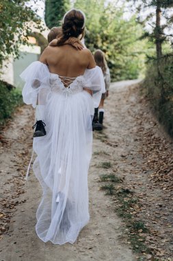 Beyaz bohem bir elbise giymiş güzel bir kadının arkadan görünüşü küçük kızını taşıyor ve yolda yürüyor. 