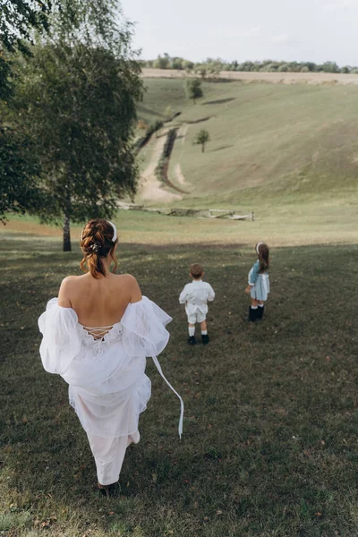 Beyaz bohem elbiseli güzel bir kadın çocuklarıyla kırsalda yürüyor. 