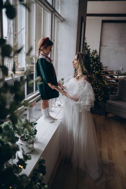 Beyaz bohem elbisesi giymiş güzel bir kadın Noel süslemeleri olan bir odada küçük kızıyla kibarca iletişim kuruyor.
