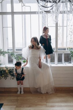 Beyaz bohem bir elbise giymiş güzel bir kadın çocukları ile birlikte noel süslemeleri olan bir odada ve oğlu annesine bir hediye veriyor ve kızı da pencerenin önünde duruyor. 