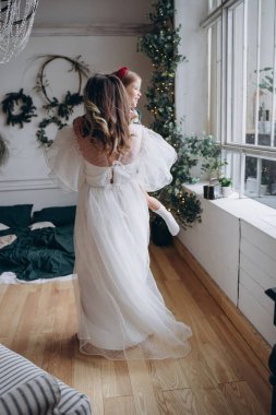 Beyaz bohem elbisesi giymiş güzel bir kadın Noel süslemeleri olan bir odada küçük kızıyla kibarca iletişim kuruyor.