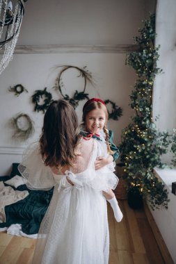 Beyaz bohem elbisesi giymiş güzel bir kadın Noel süslemeleri olan bir odada küçük kızıyla kibarca iletişim kuruyor.